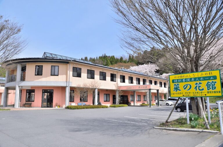 介護施設の建物外観。ベージュと赤茶色の外壁、駐車場と案内看板が見える。