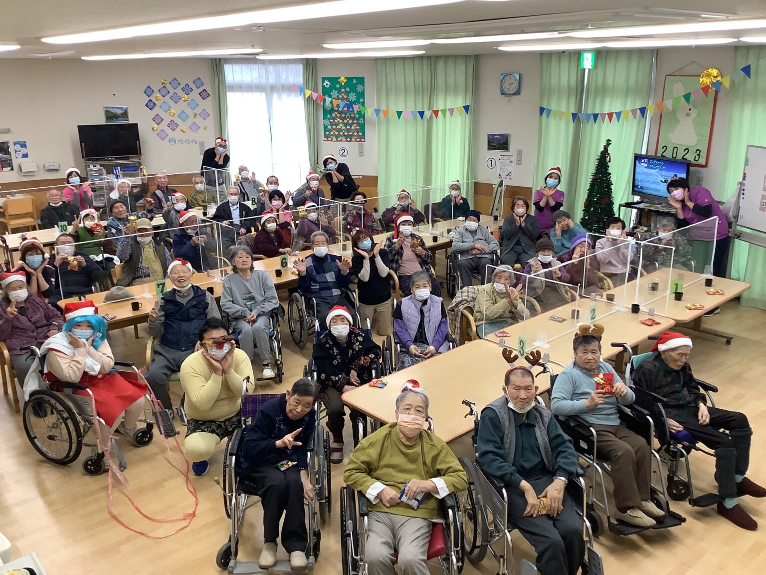 介護施設でのクリスマス行事。多くの入居者と職員が参加し、装飾された共用スペースで楽しい時間を過ごしている。