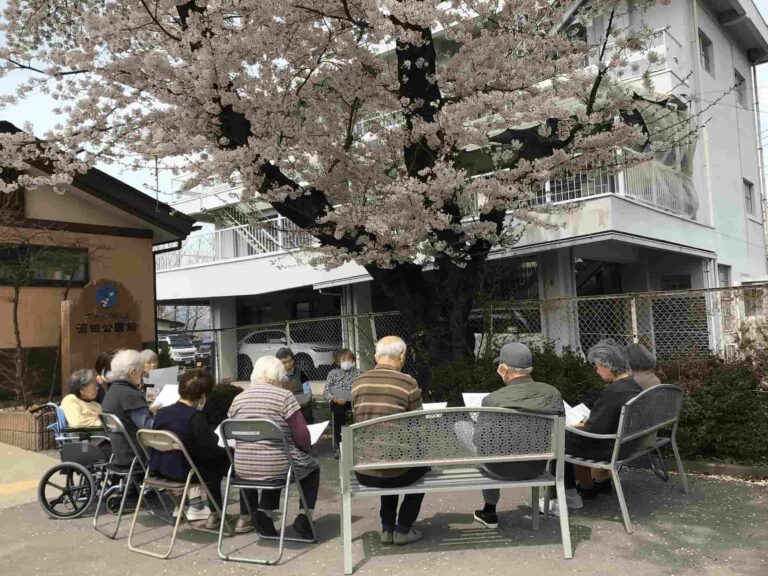 桜の下で入居者たちが談笑する様子。施設の庭での春の風情のある一場面。